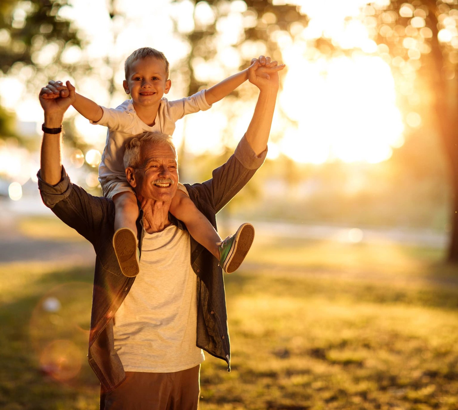 A young boy sits on top of his grandfather's shoulders, both smiling.