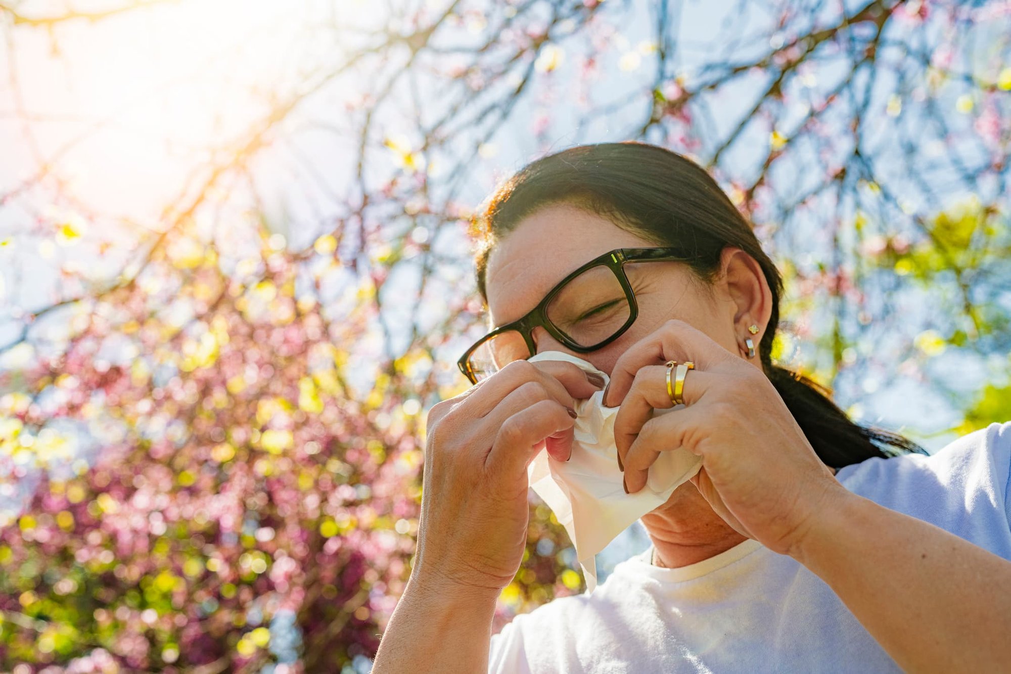 Woman experiencing seasonal allergy symptoms outdoors during spring pollen season while sneezing into a tissue